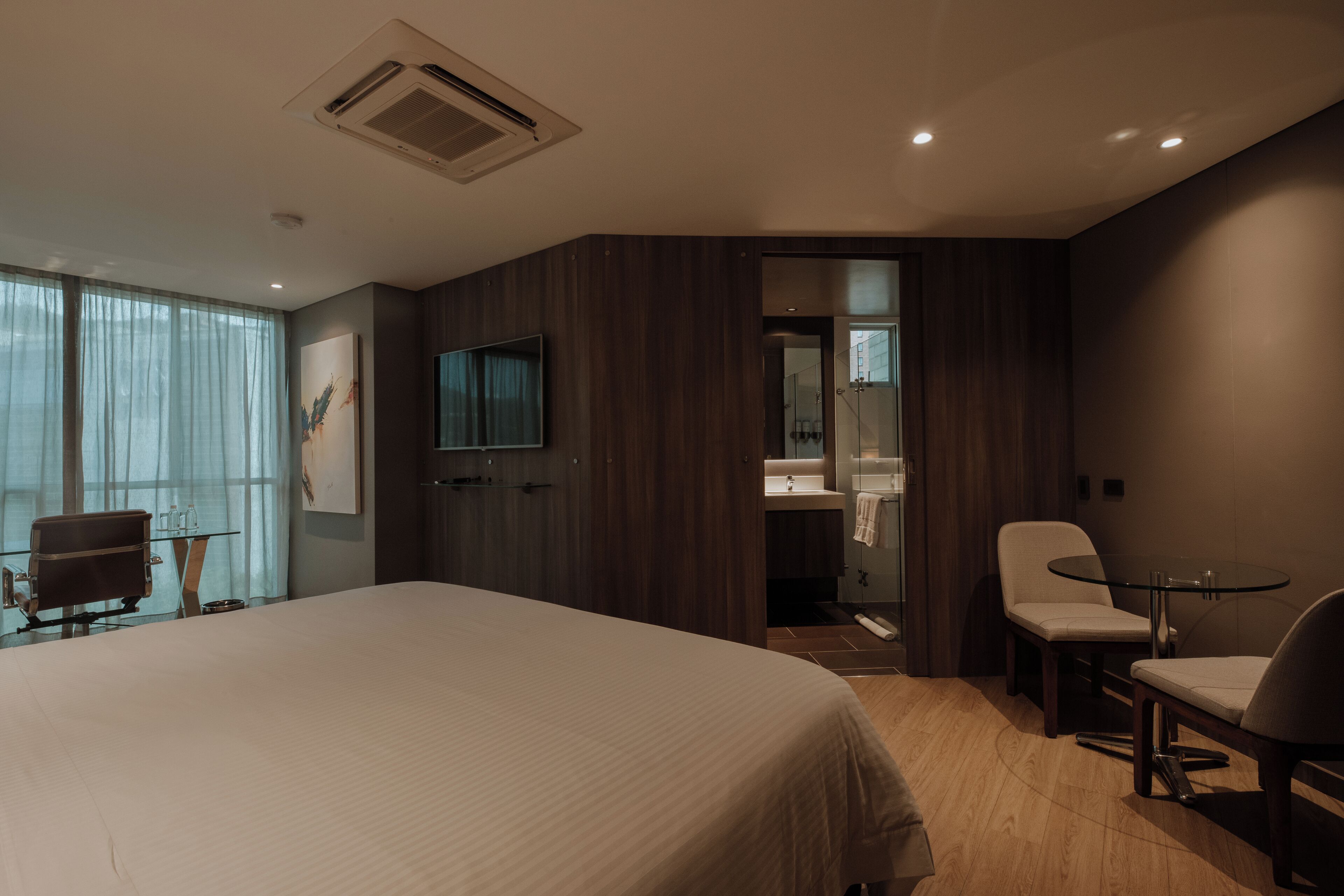 A modern hotel room with a large bed, a seating area with a table and chairs, a television, and a bathroom visible through an open door.