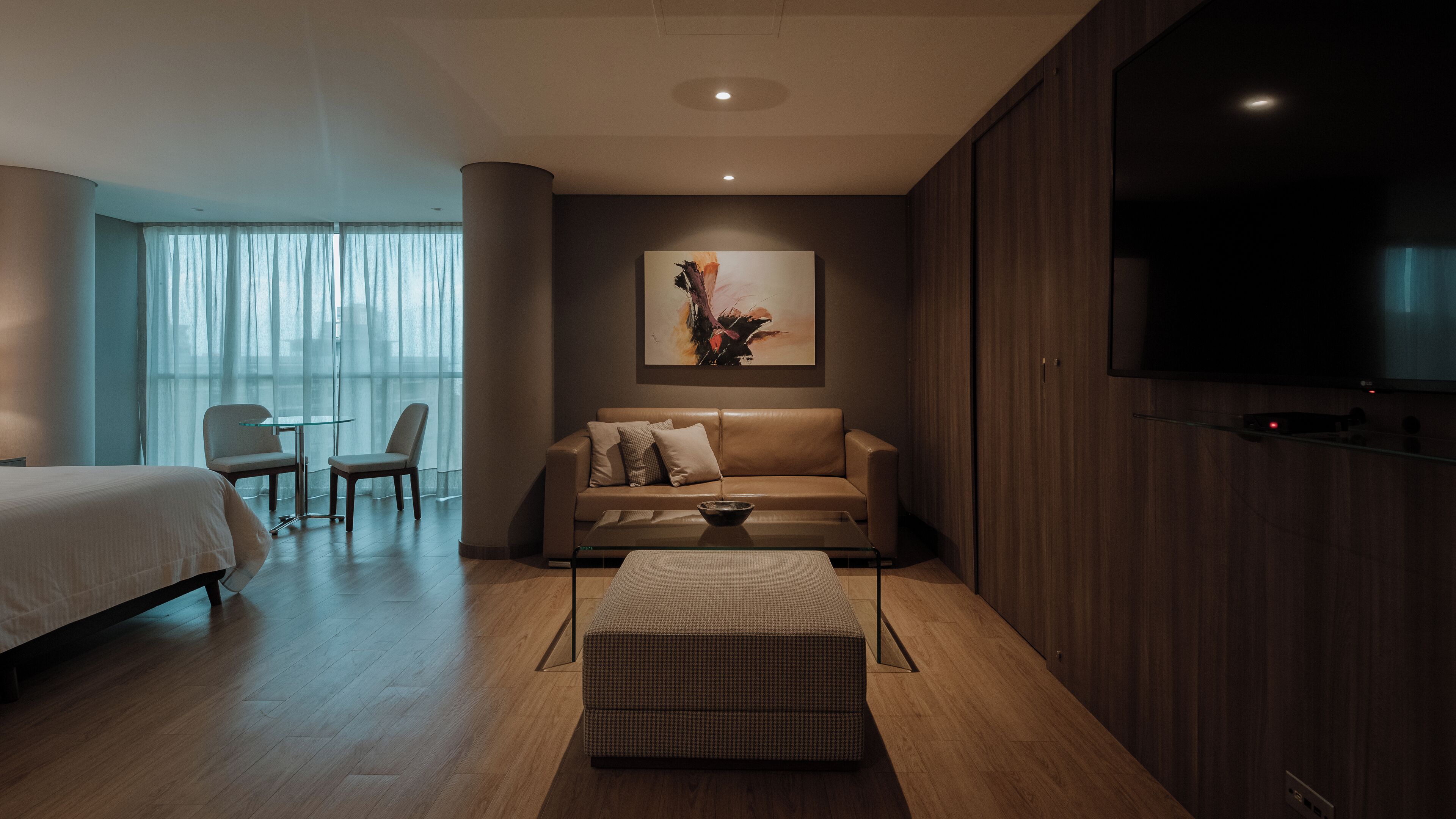 Quarto de hotel moderno com cama, sofá, mesa de jantar com cadeiras, televisão e uma grande janela com cortinas translúcidas.