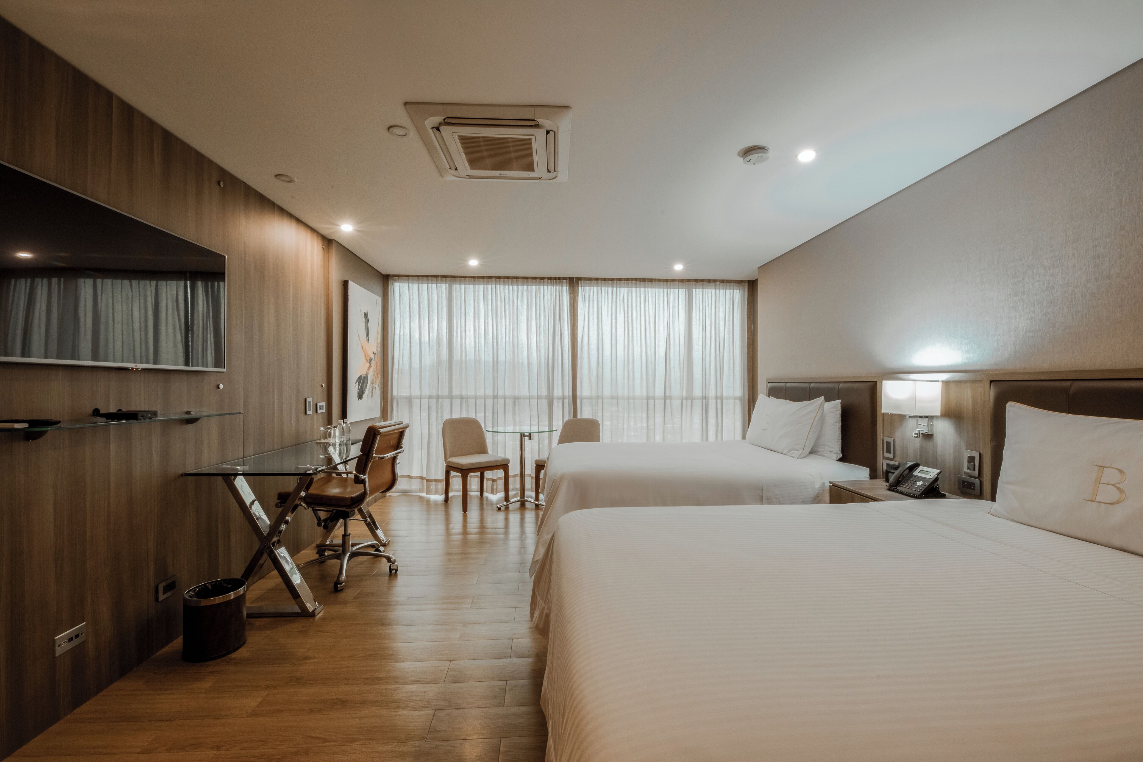 A hotel room with two beds, a desk, a chair, a small table, and a TV mounted on the wall.