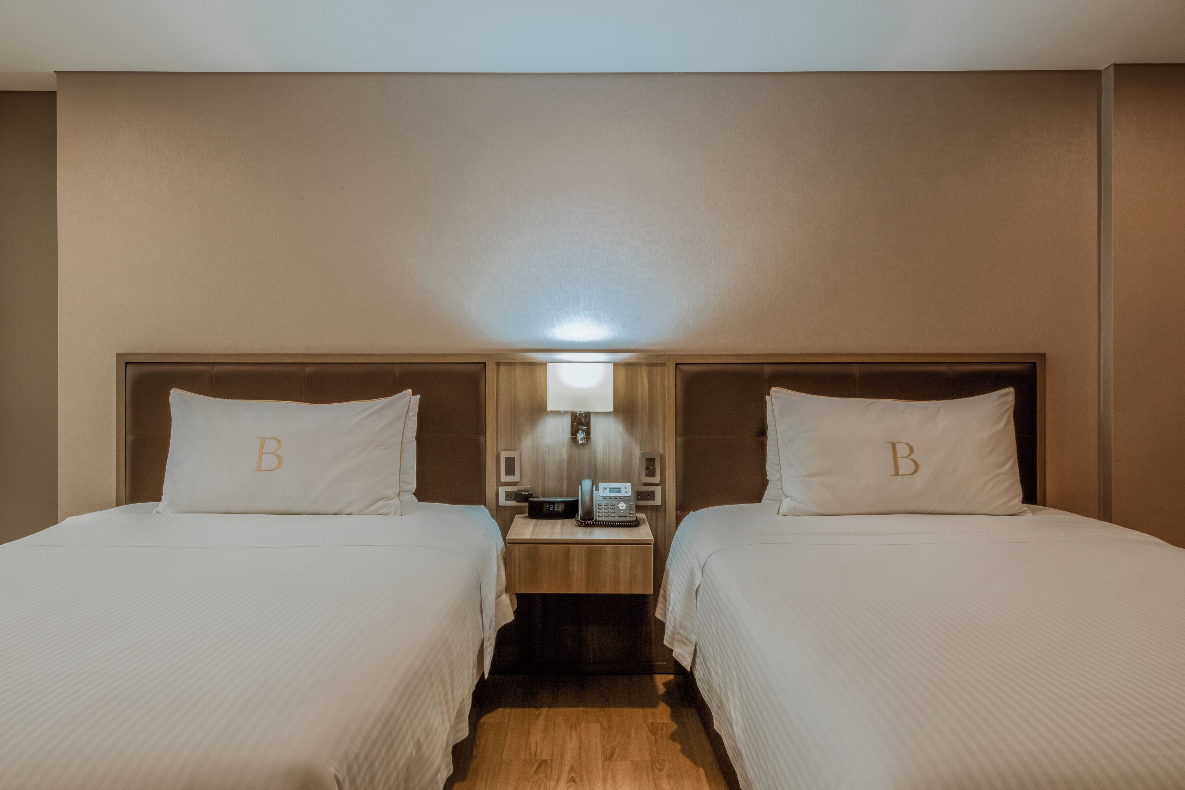 Ein Hotelzimmer mit zwei Einzelbetten, jedes mit einem Kopfkissen, das mit einem 'B' gekennzeichnet ist. Es gibt einen Nachttisch mit einer Lampe und einem Telefon.