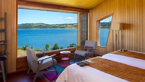 A bedroom with a large window offering a view of a lake and hills. There is a bed, two chairs, and a small table. - Refugia Chiloé (Castro)