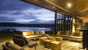 A spacious outdoor seating area with multiple sofas and ottomans, large windows offering a view of a lake at dusk. - Refugia Chiloé (Castro)