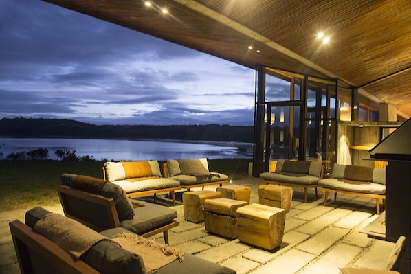 Un amplio espacio al aire libre con varios sofás y otomanas, grandes ventanales que dan a una vista del lago al anochecer.