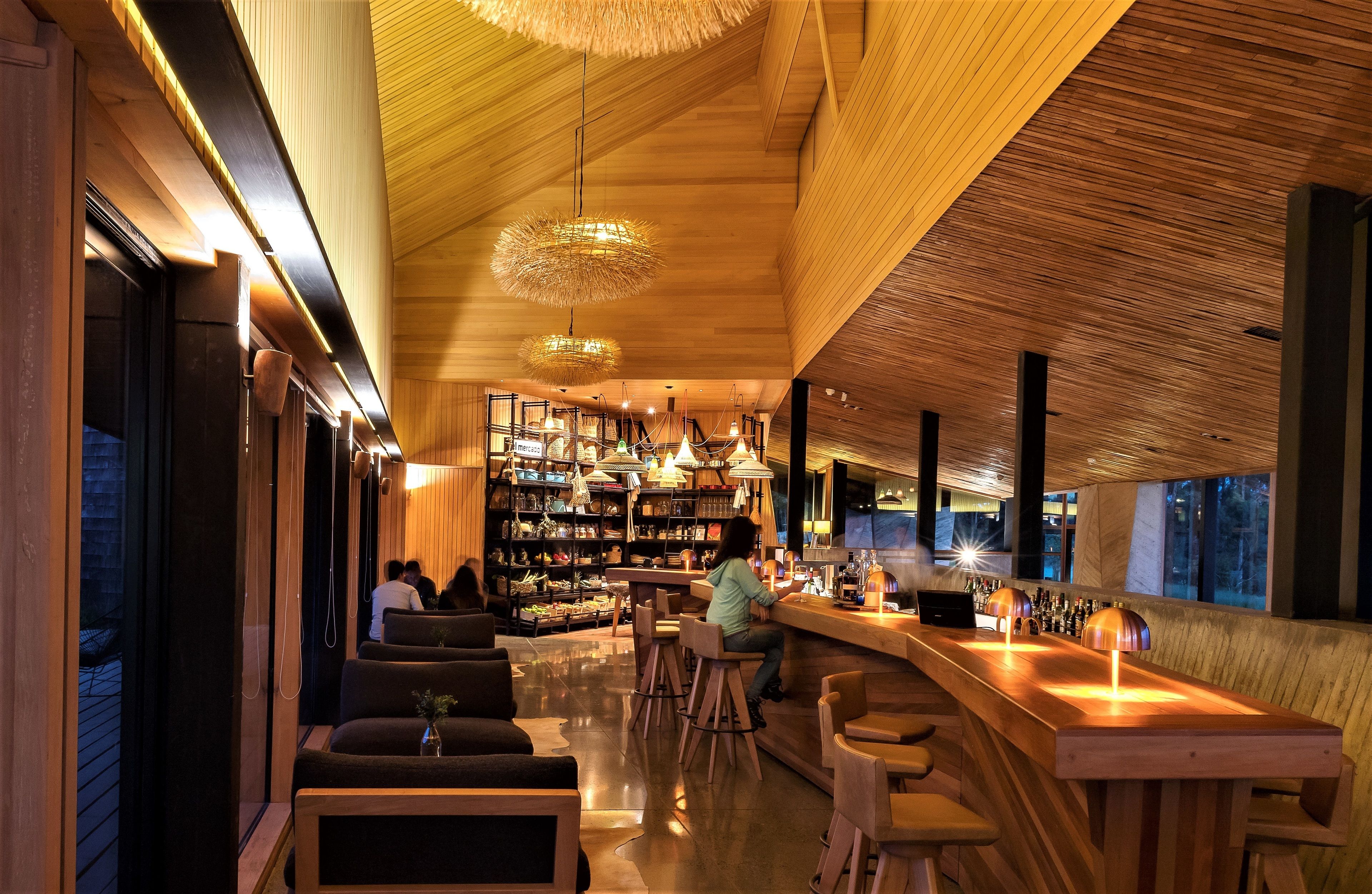 Zona de bar moderna con mobiliario de madera, lámparas colgantes y una barra repleta de botellas.