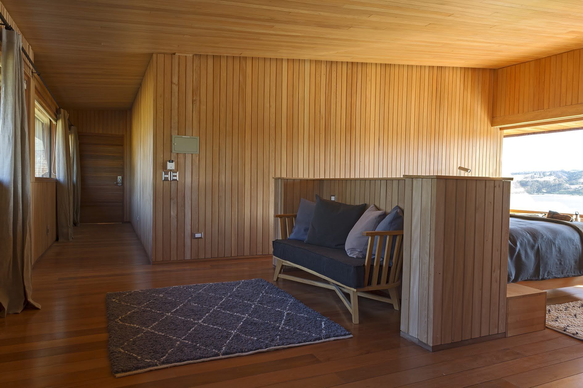 Interior de una cabaña de madera con un sofá cama, una alfombra y una ventana con vistas.