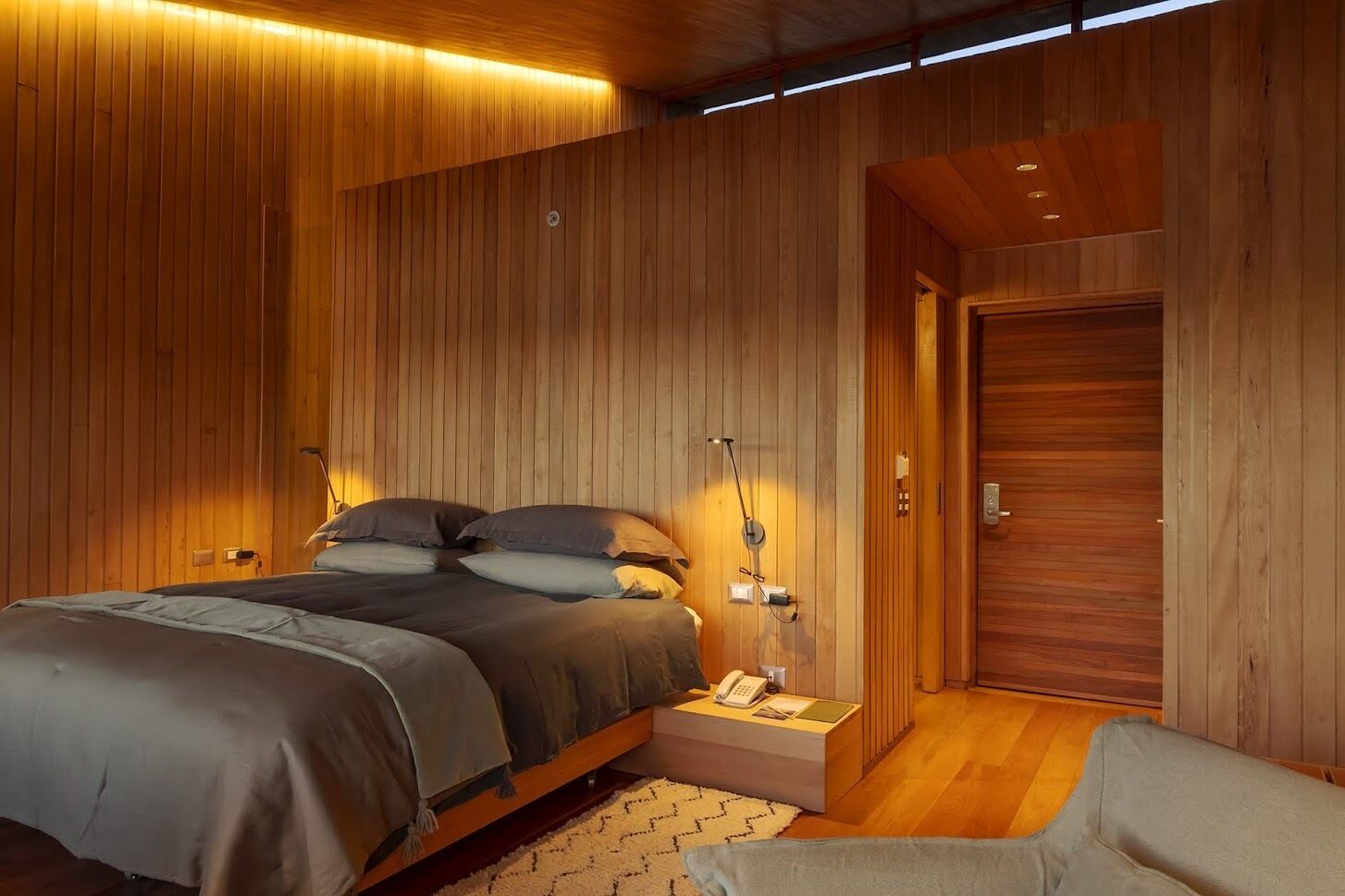 Ein modernes Schlafzimmer mit einer Holzvertäfelung, einem großen Bett mit Kopfteil, Nachttischlampen und einem kleinen Nachttisch mit einem Telefon.