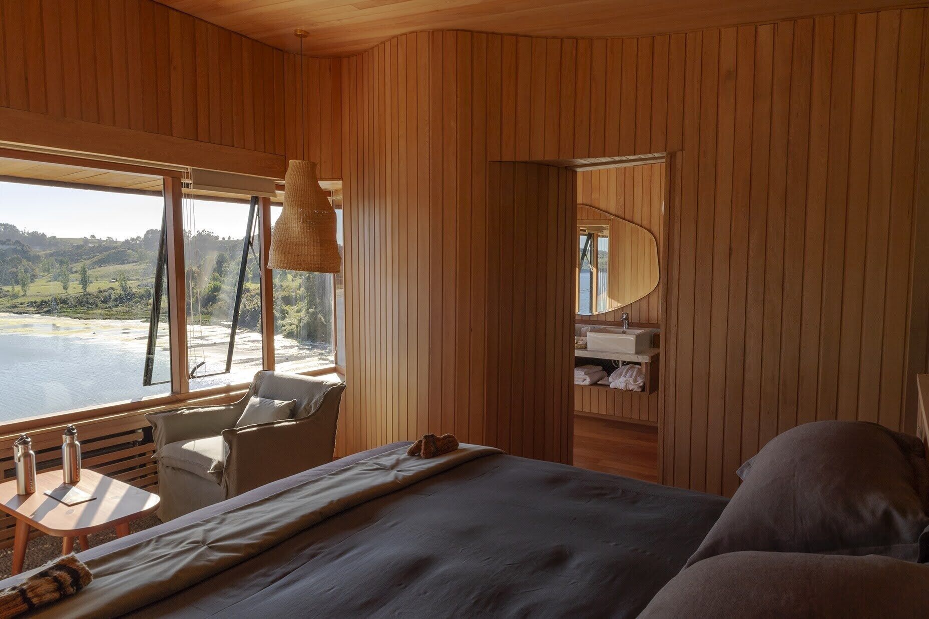 Una cabaña de madera con una cama, una mesita con botellas, una silla y vistas a un lago.