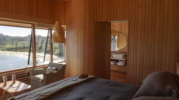 Una cabaña de madera con una cama, una mesita con botellas, una silla y vistas a un lago.