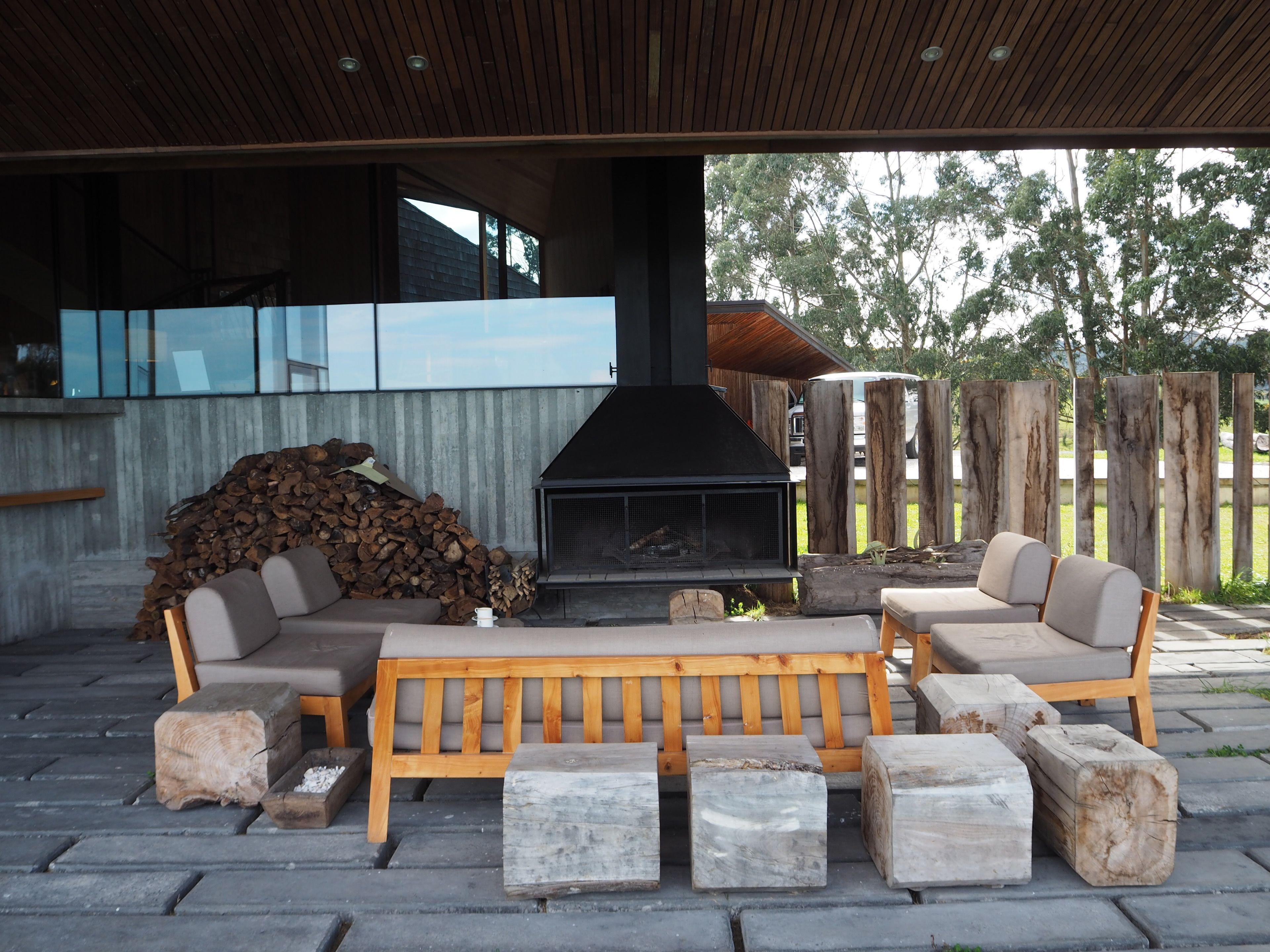 An outdoor seating area with a built-in fireplace, wooden furniture, and a stack of firewood.
