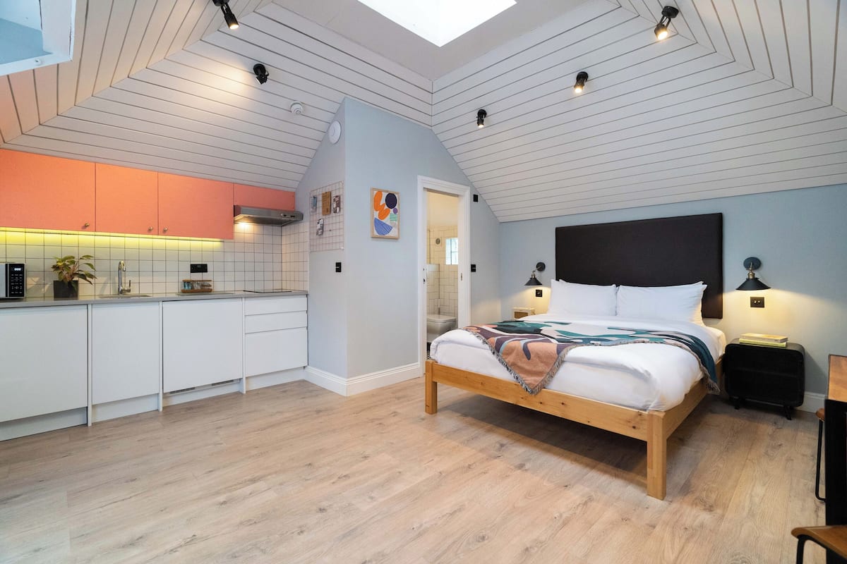 A modern loft bedroom with a skylight, a wooden bed, a nightstand, and a kitchen area with white cabinets.