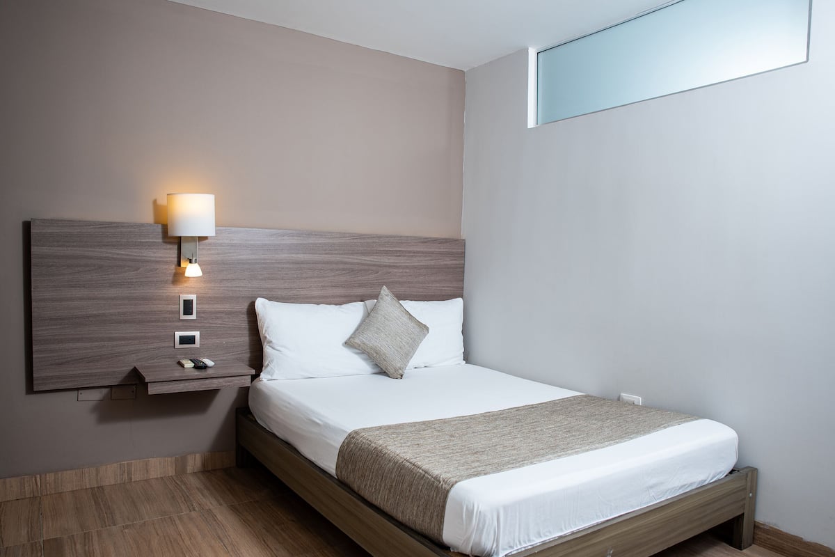Un lit bien fait, avec une tête de lit en bois, une lampe de chevet et une petite étagère sur laquelle repose une télécommande.