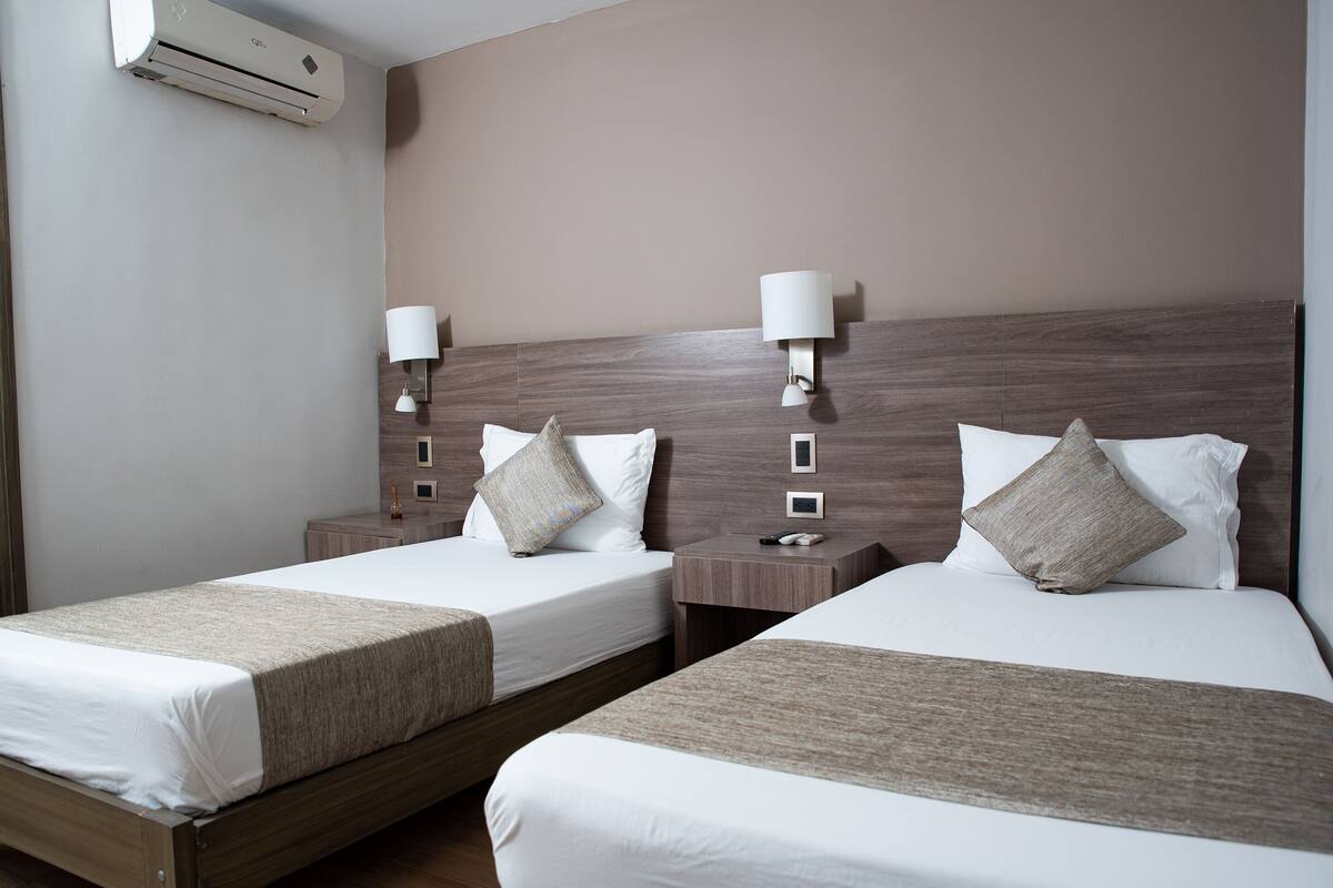 Une chambre d’hôtel avec deux lits simples, chacun doté d’une tête de lit en bois, de draps blancs et d’un plaid beige. On y trouve des tables de chevet avec des lampes et un climatiseur fixé au mur.