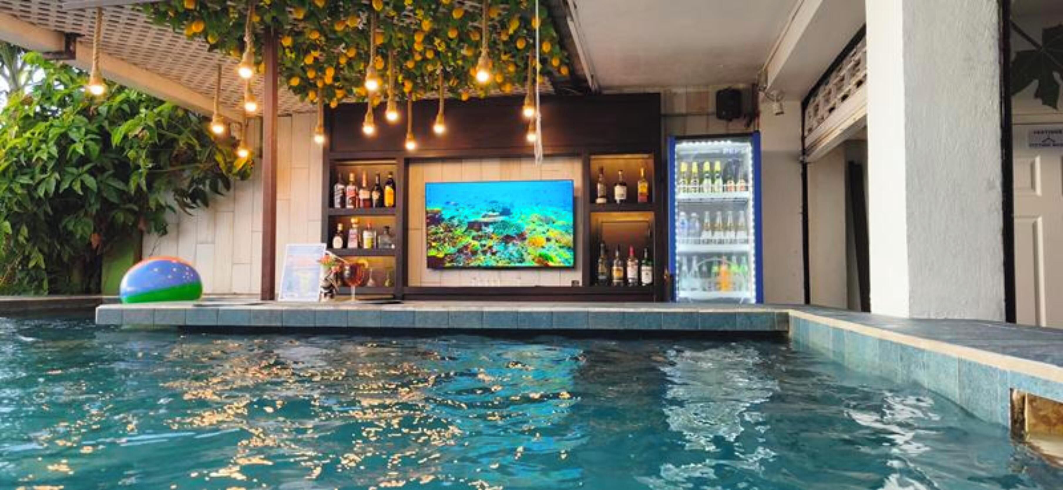 A swimming pool with a bar area, including a television screen, shelves with bottles, and a refrigerator.
