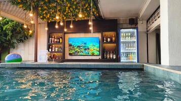 A swimming pool with a bar area, including a television screen, shelves with bottles, and a refrigerator.