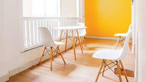 A bright room with wooden ceiling beams, white walls, and a yellow accent wall. There are four white chairs and a small table.