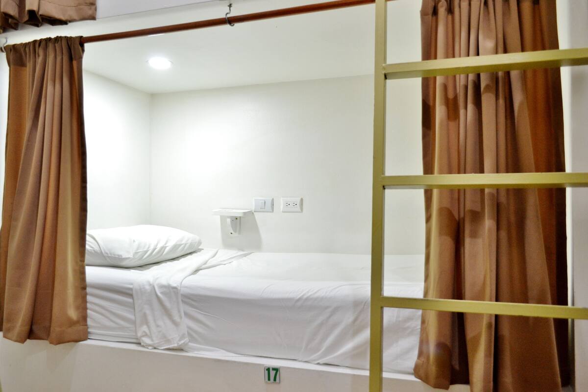 A single-bed room with a white bed, brown curtains, a white wall, and a small white table.