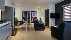 Un salon moderne avec un canapé noir, un téléviseur fixé au mur et un coin cuisine équipé d’un réfrigérateur.