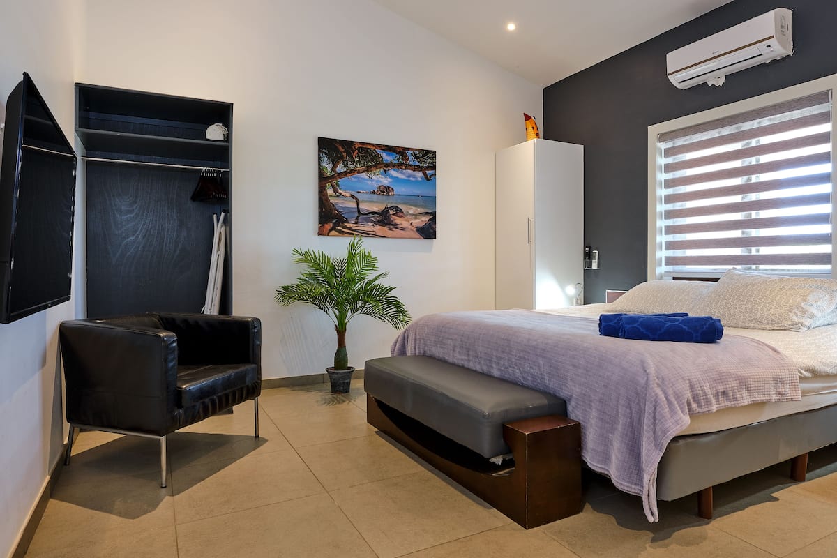 Ein modernes Schlafzimmer mit Bett, einer Bank, einer Topfpflanze, einem Kleiderschrank und einem Gemälde an der Wand.