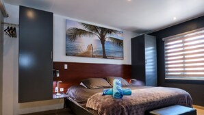 Une chambre moderne avec un grand lit, une table de chevet, un bureau, un tableau représentant une plage accroché au mur et une fenêtre avec des stores.