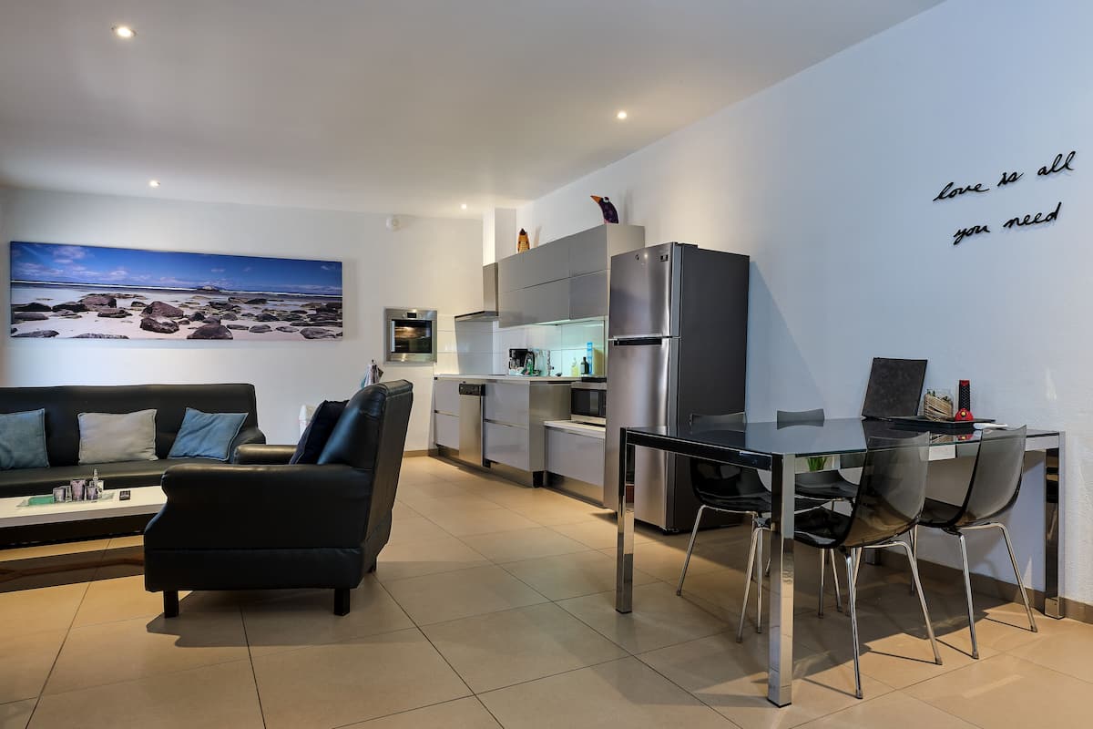 Ein modernes Wohnzimmer mit einem schwarzen Ledersofa, einem Glastisch mit Stühlen, einer Küchenzeile mit Edelstahlgeräten und einem großen Gemälde, das eine Strandlandschaft zeigt.