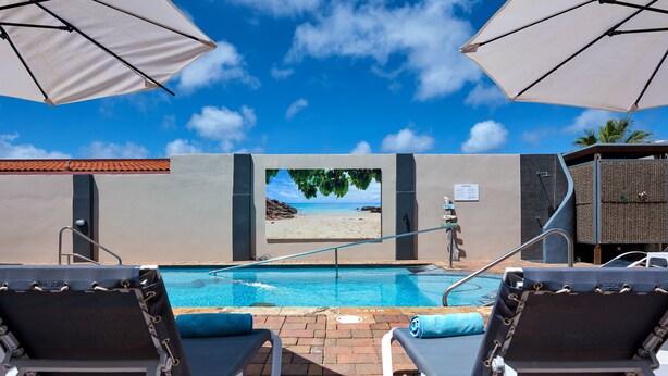 Un espace aménagé au bord de la piscine, avec des chaises longues, des parasols et une vue sur la plage, visible à travers une fenêtre.