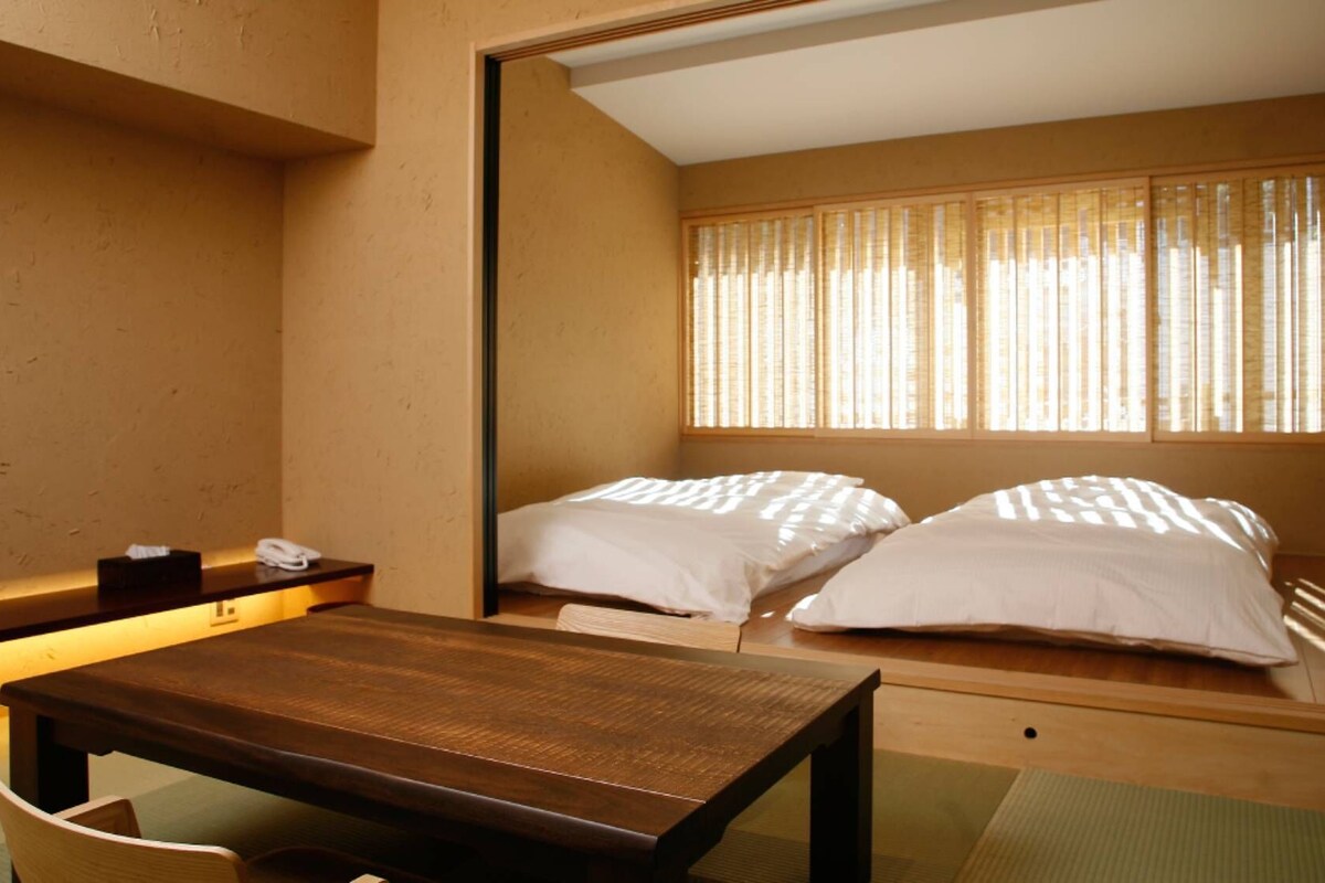 A hotel room with a wooden table, two beds, a mirror, and a window with blinds.