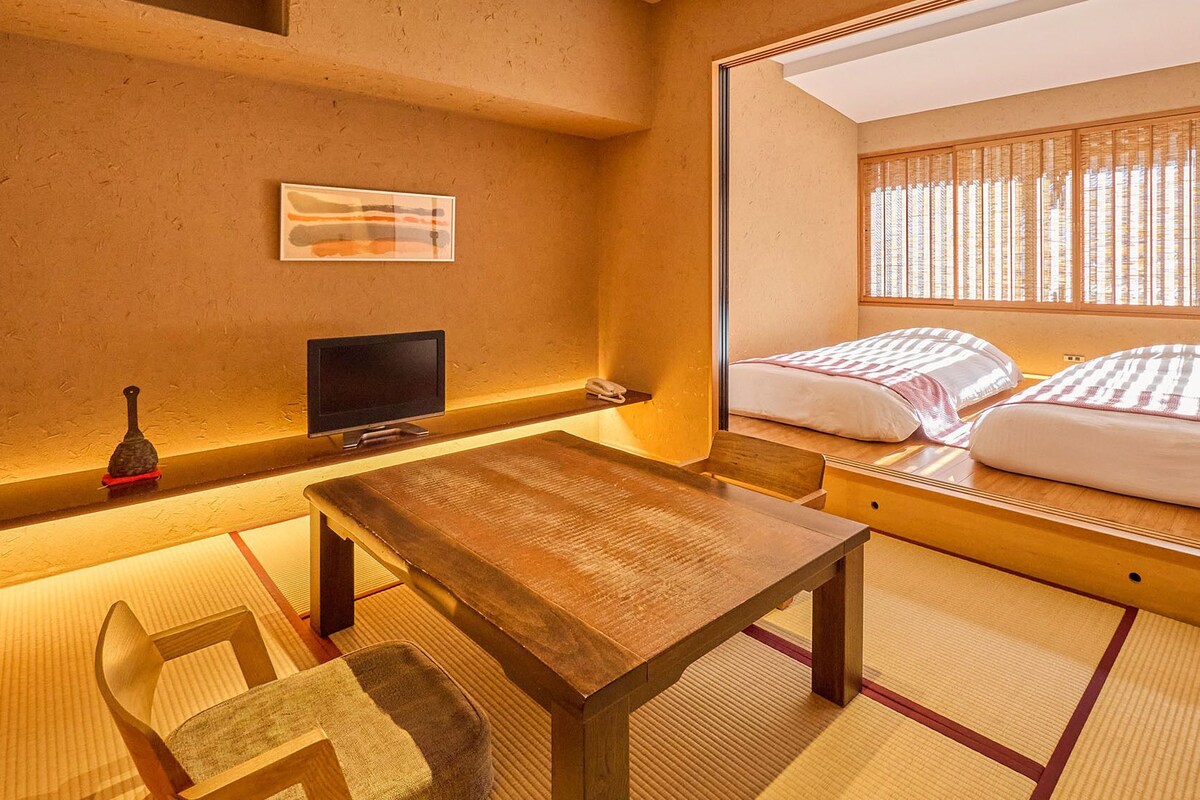 A traditional Japanese room with tatami flooring, a wooden table, a television, and a bed with a view of the outside through a window with shoji screens.