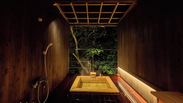 Una sauna de fusta amb una banyera hidromassatge rectangular, una zona de dutxes i vista a arbres a través d'un separador de vidre.