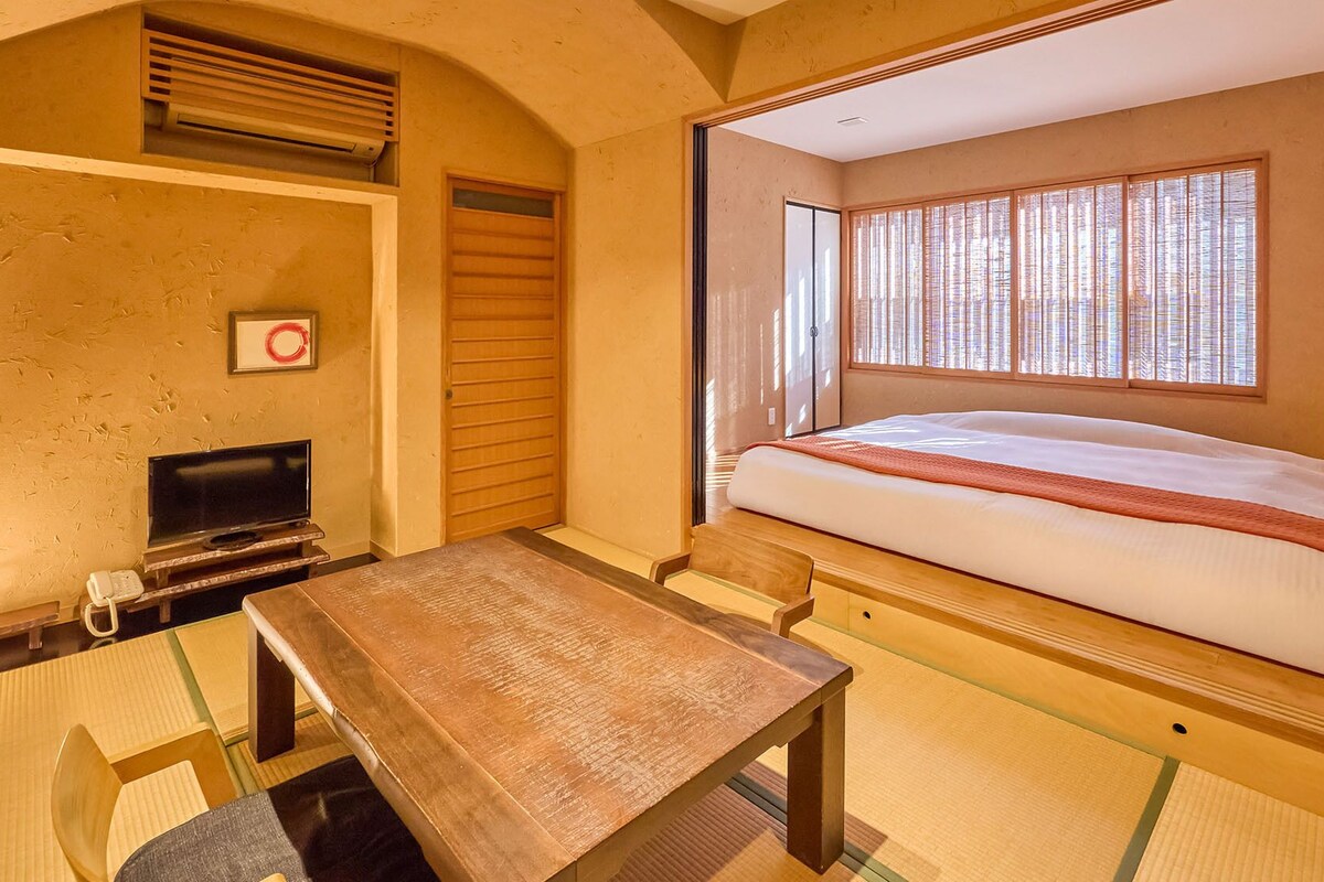 A room with a bed, a television, a table, and a window with blinds.