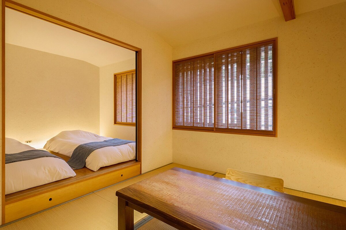 A room with a bed, a wooden table, and a window with vertical blinds.