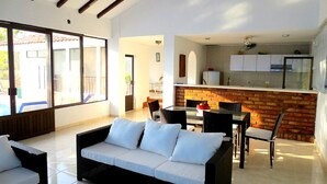 Amplia sala de estar con sofá, sillas y una mesa de comedor. Cuenta con una zona de cocina con salpicadero de ladrillo y armarios.