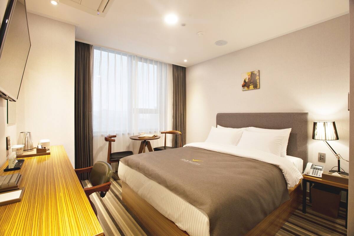 A hotel room with a large bed, a desk with a computer, a chair, a small table, a lamp, and a painting on the wall.