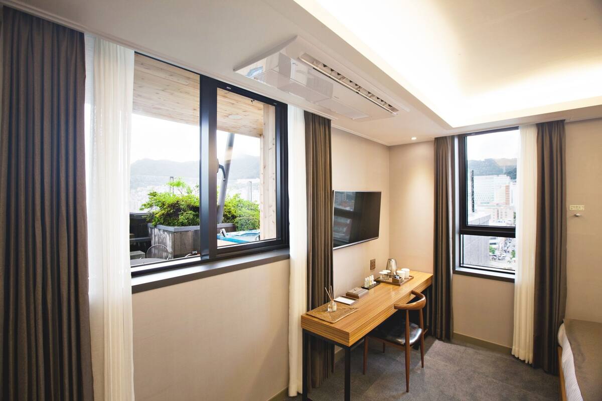 A hotel room with a wooden desk, a flat-screen TV, and large windows offering a city view.
