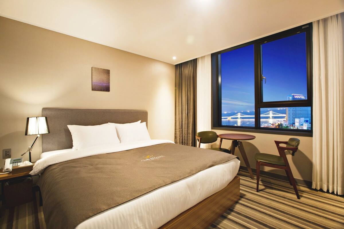 A hotel room with a large bed, a desk with a lamp, a chair, and a window with a city view.