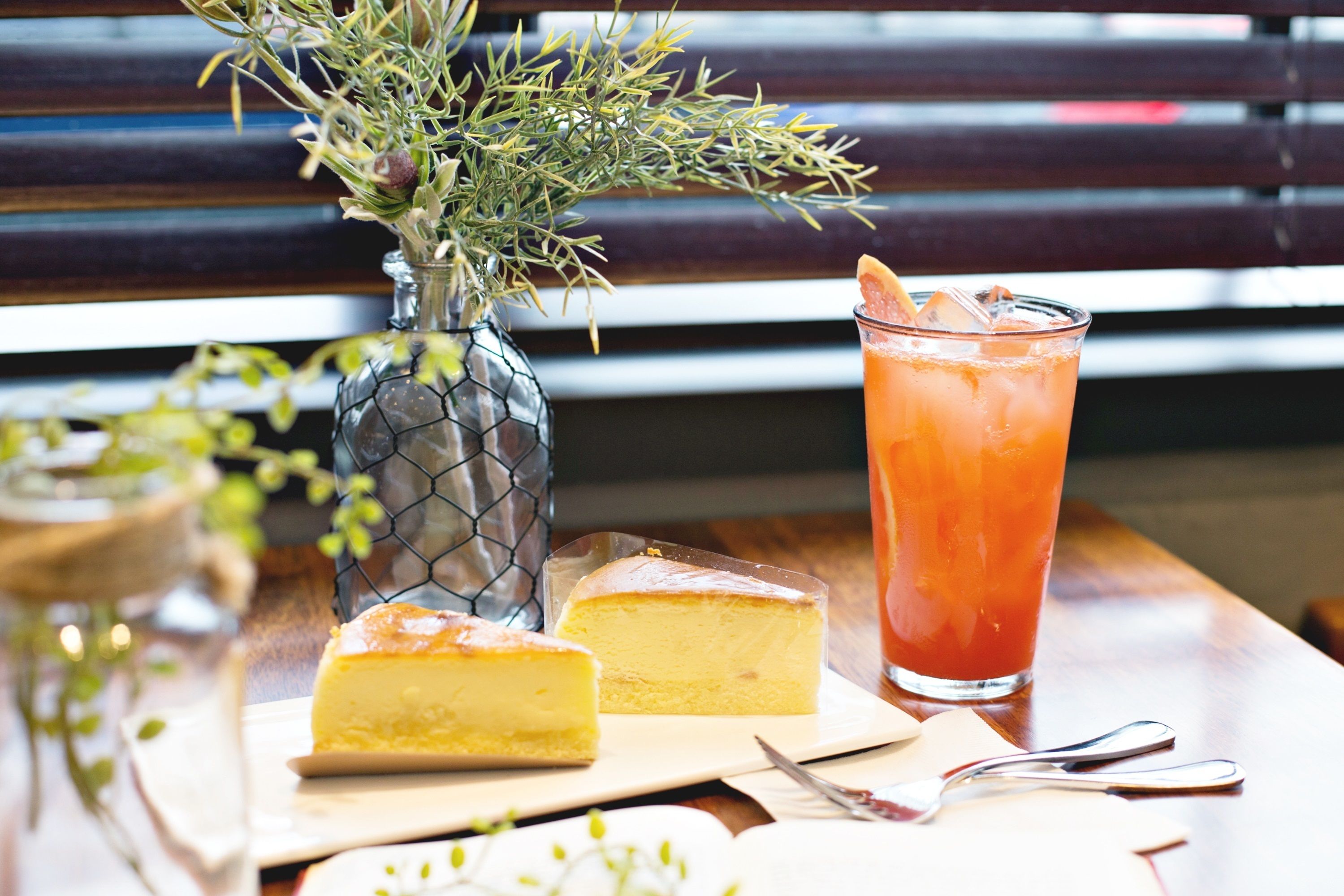 A table with a glass of iced drink, a slice of cheesecake, and a decorative vase with greenery.