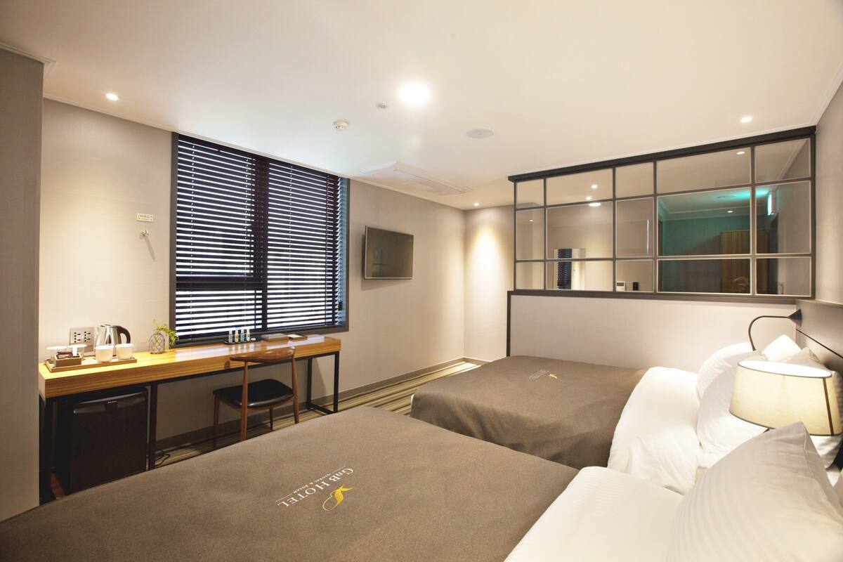 A hotel room with two beds, a desk, a chair, a TV, and a window with blinds.