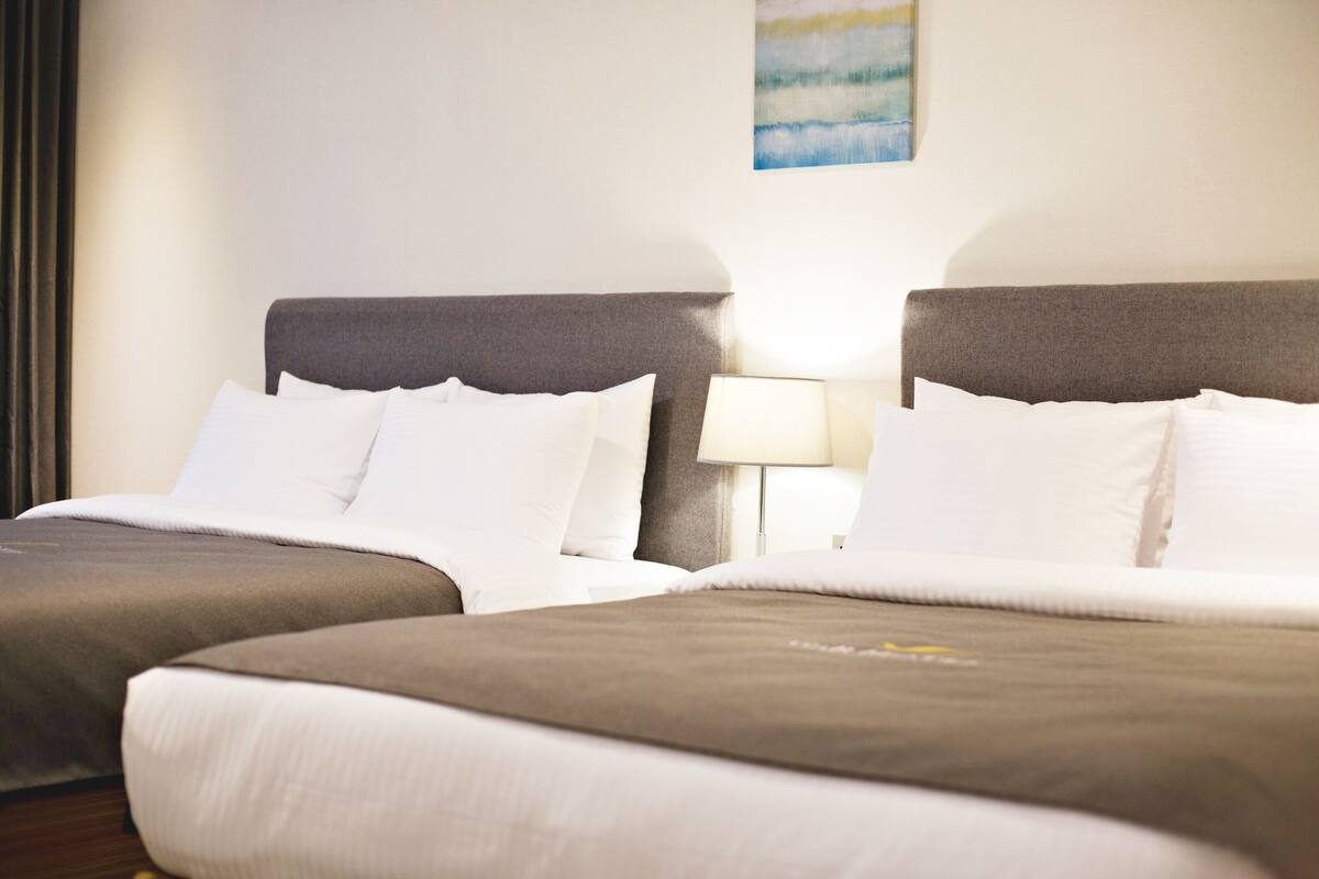 A hotel room with two beds, each with a grey headboard, white bedding, and a bedside lamp.