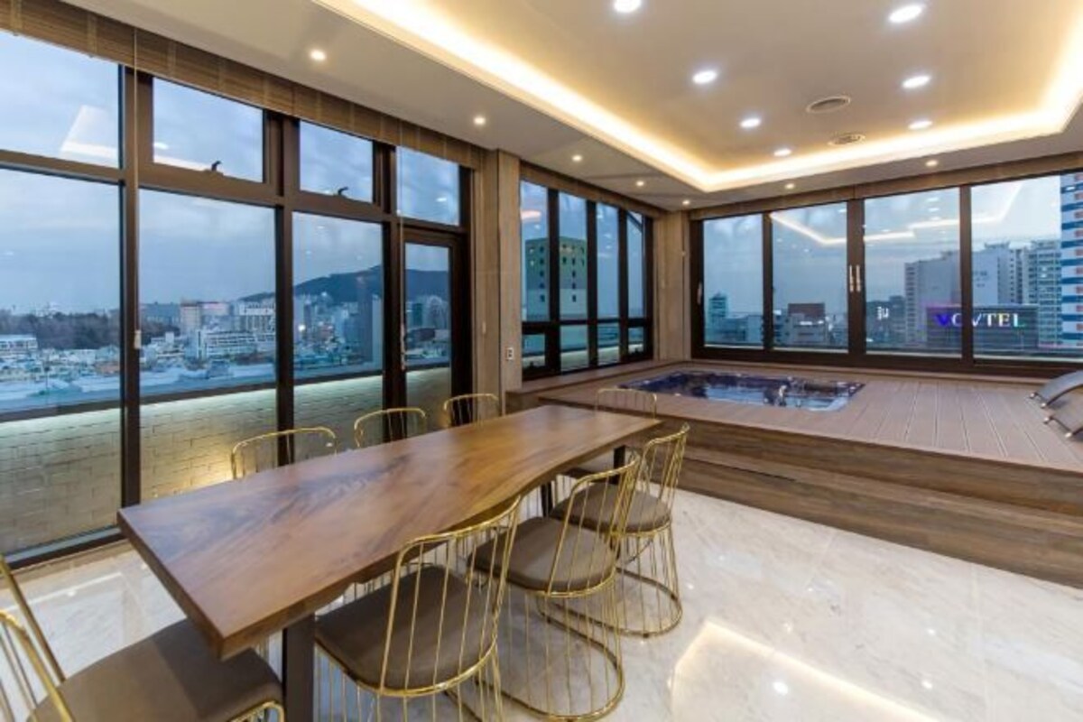 A modern hotel room with a large wooden dining table, comfortable chairs, and a hot tub.