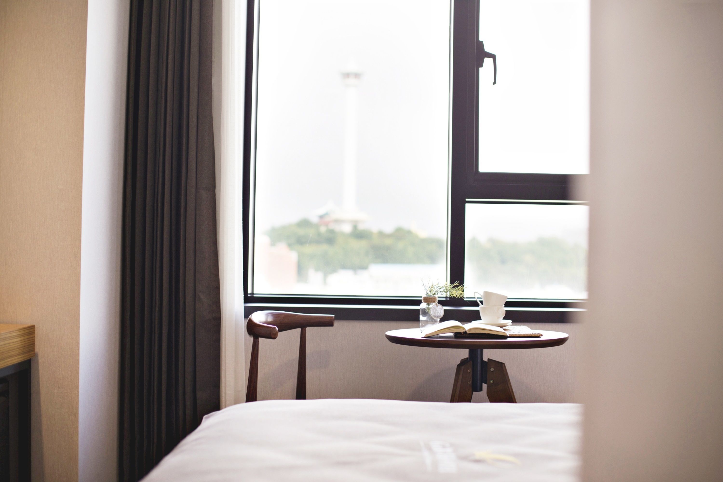 Une chambre d’hôtel avec un lit, une petite table sur laquelle se trouvent un livre et une tasse, une chaise et une fenêtre donnant sur un grand bâtiment et de la verdure.