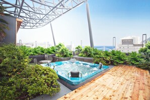 A rooftop patio with a hot tub, wooden deck, and lush greenery.