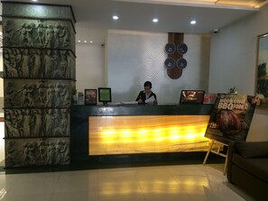 Una recepción de hotel con una recepcionista, una gran escultura de mármol, un reloj y un cartel anunciando una cena de barbacoa de fin de semana.