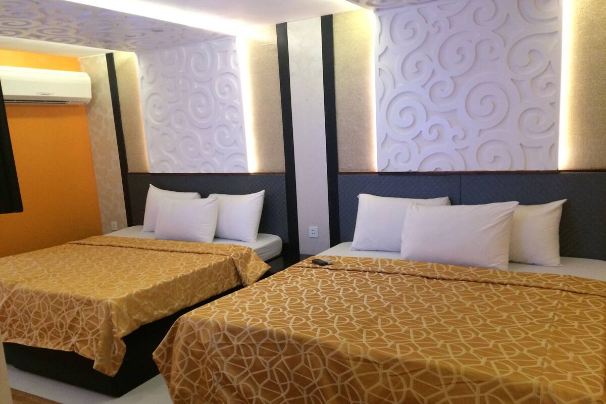 A hotel room with two single beds, each with a patterned bedspread and white pillows. The room has a modern design with decorative wall panels and a built-in air conditioning unit.