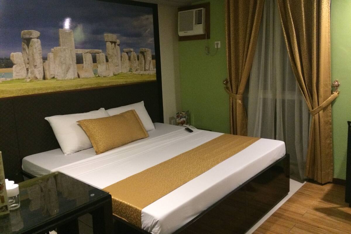 A hotel room with a bed, a nightstand, a painting of Stonehenge, a green wall, and a window with curtains.