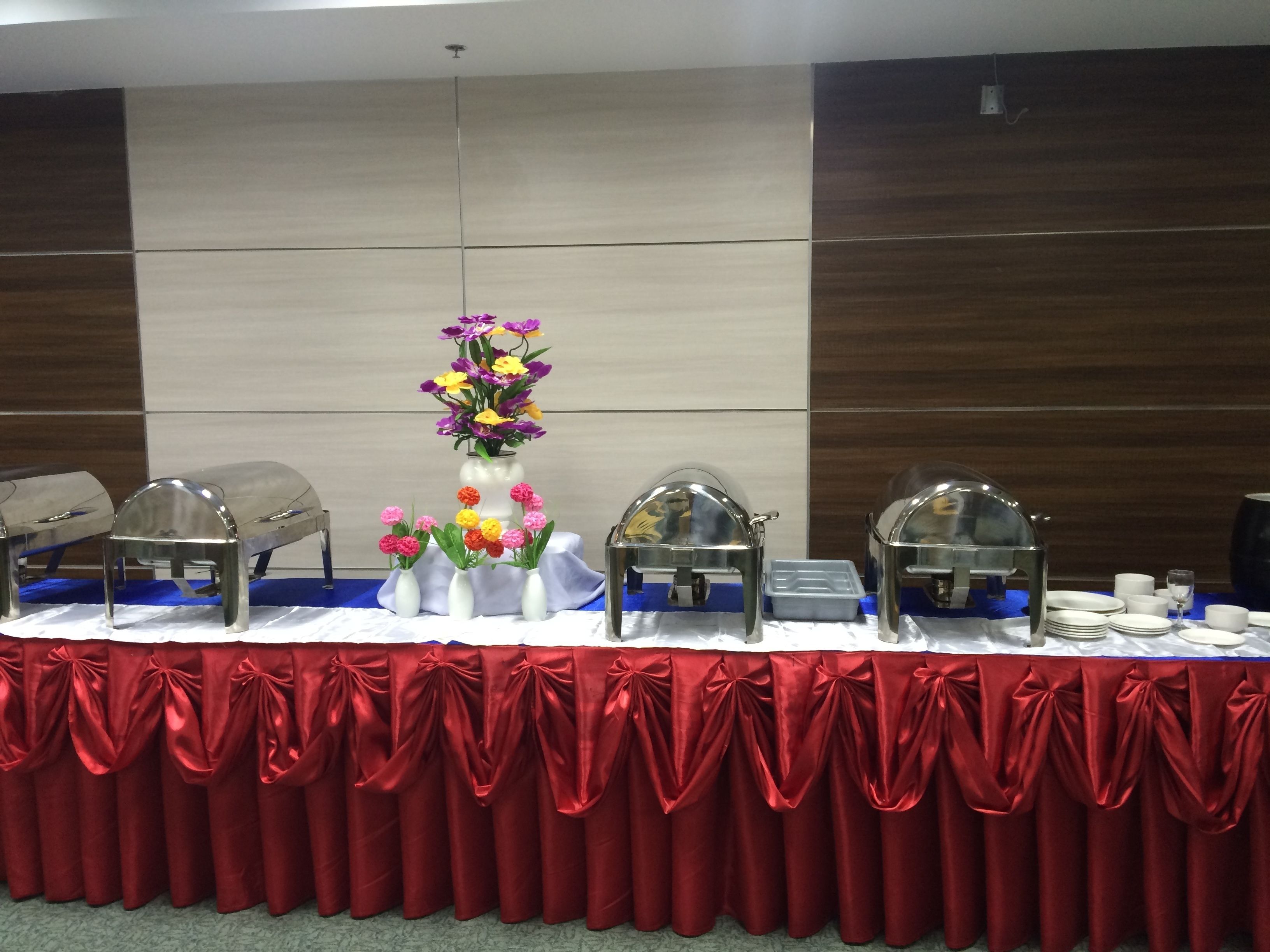 A buffet table with food service stations, a central floral arrangement, and a red draped tablecloth.