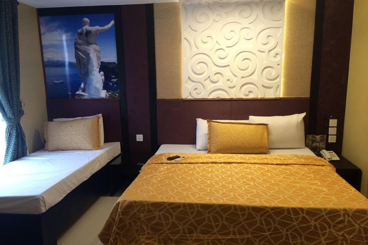 A hotel room with two beds, one with a patterned bedspread and the other with plain sheets. There is a decorative wall panel and a painting of a figure on the wall.