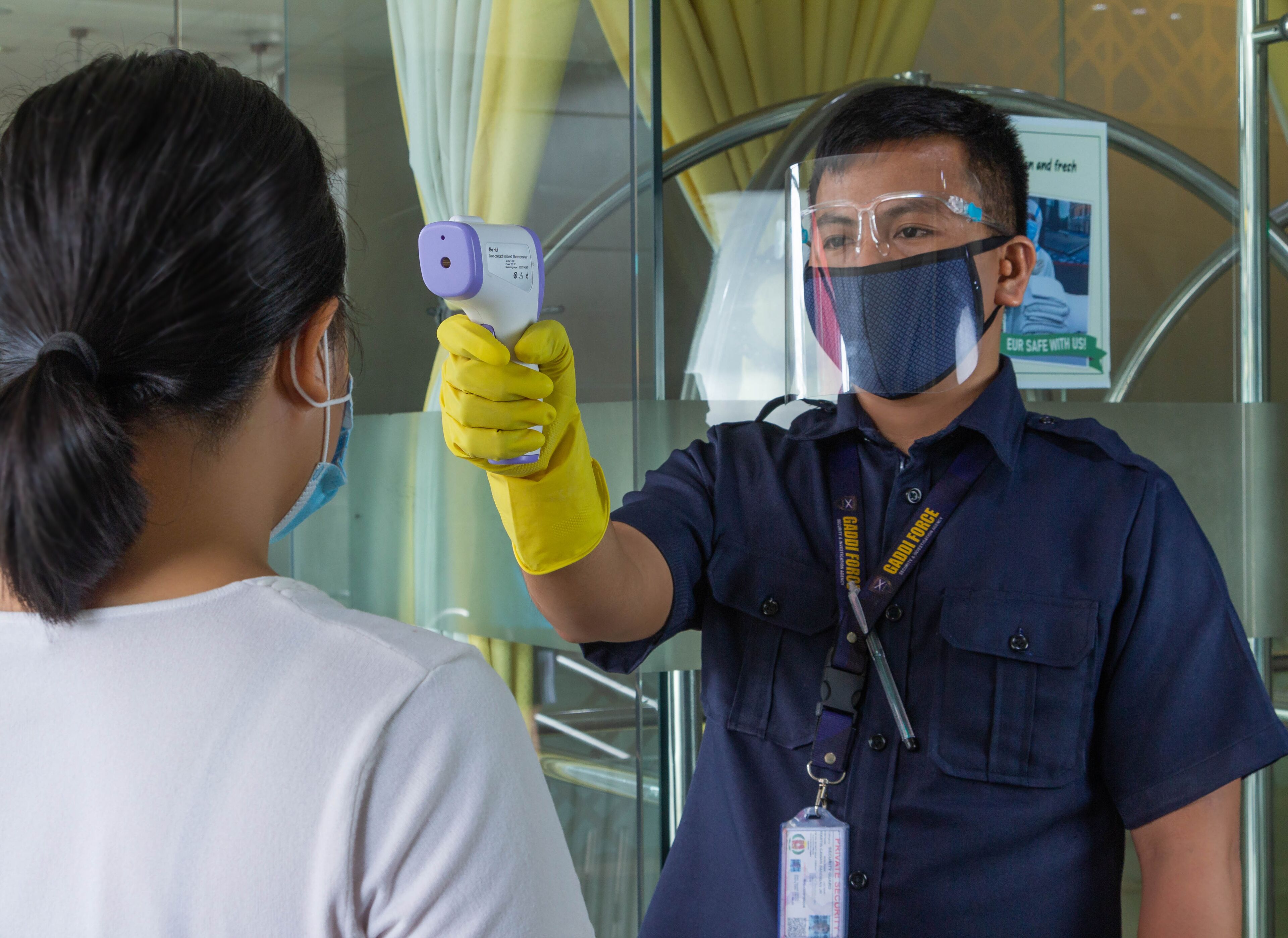 A person wearing a blue uniform and yellow gloves holding a handheld device, likely for temperature screening.