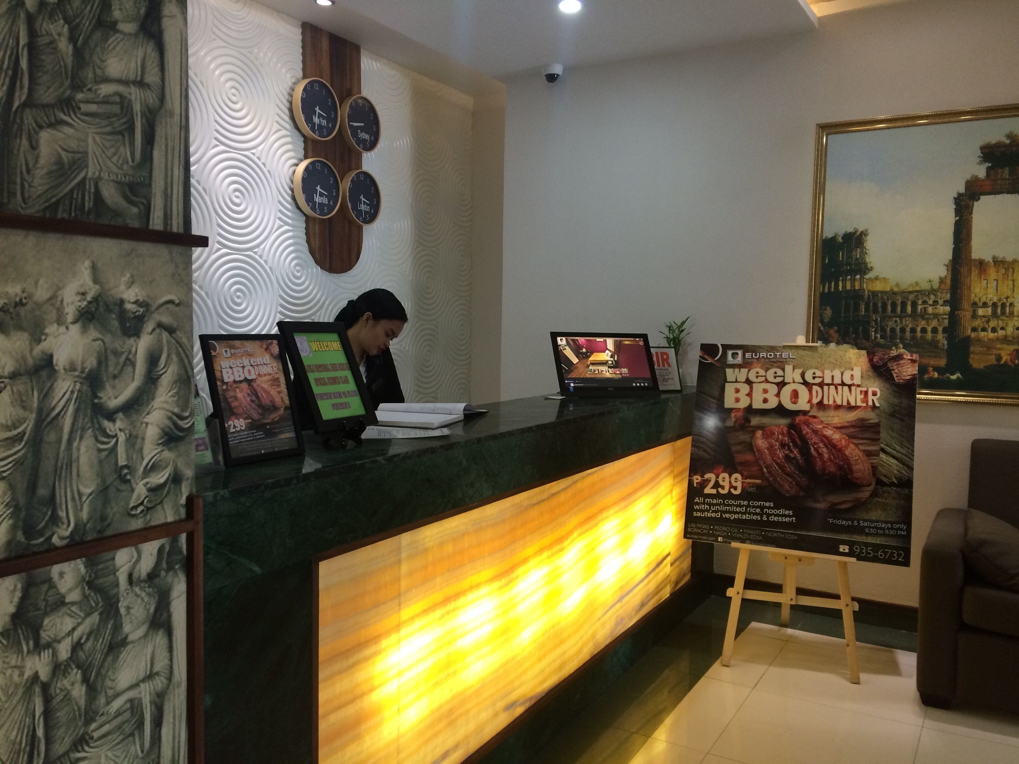 A hotel reception desk with a digital screen displaying a welcome message, a poster for weekend BBQ dinner, and a receptionist.