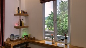 Una habitación con un escritorio de madera, estanterías con botellas y vasos, una ventana con vista a la vegetación y un comedor con sillas.