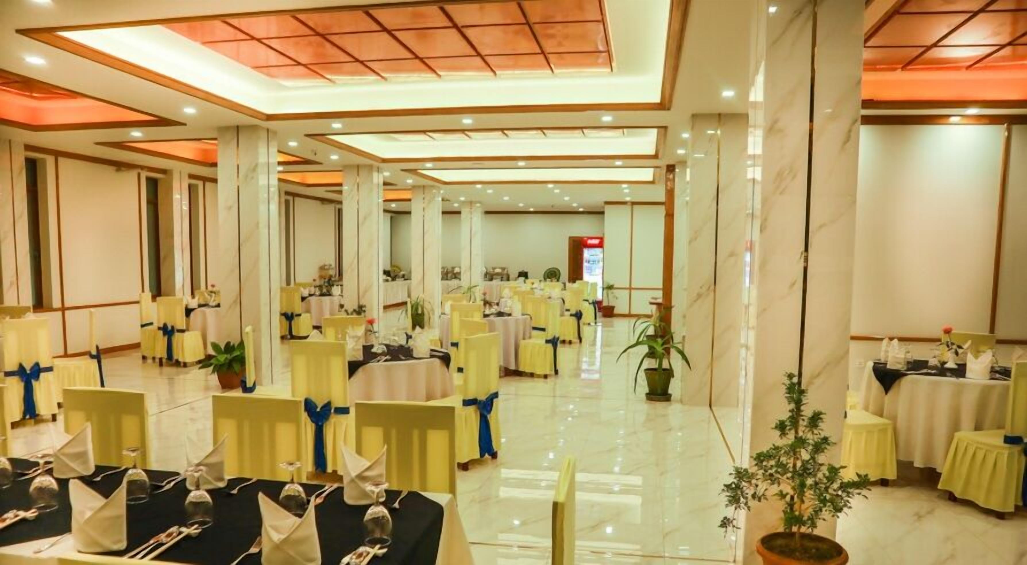 Un amplio salón de banquetes con varias mesas redondas, cada una con mantel blanco y lazadas azules. La sala tiene un techo alto con paneles de madera e iluminación empotrada.