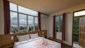 Habitación de hotel con una cama grande, cabecera de madera, una mesita de noche con teléfono, un banco y vistas de la ciudad a través de amplias ventanas.
